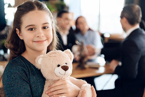 Young girl smiling and holding a teddy bear while parents talk with a lawyer
